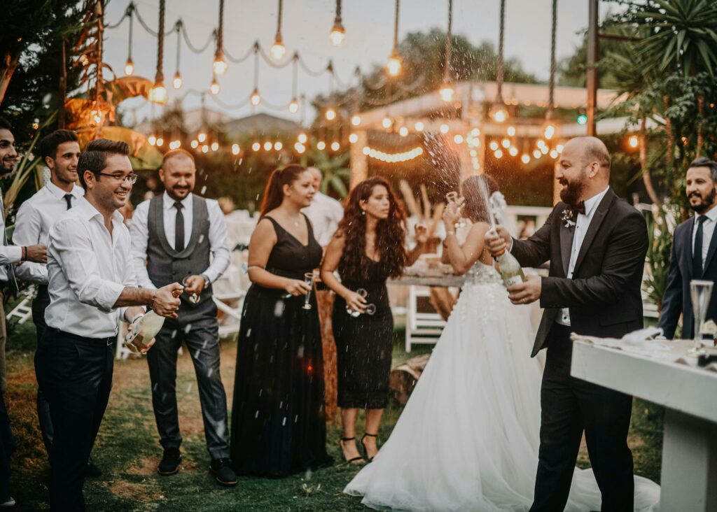 Joyful wedding party celebration outdoors with champagne and smiling guests dressed in formal attire.